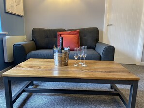 A wooden coffee table with a metal bucket containing a bottle and glasses, placed in front of a grey sofa with a red pillow.