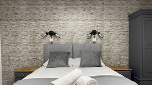 A neatly made bed with a grey blanket and pillows, a white towel, and a brick-patterned wall behind. - The Roade House Rooms (Northampton)