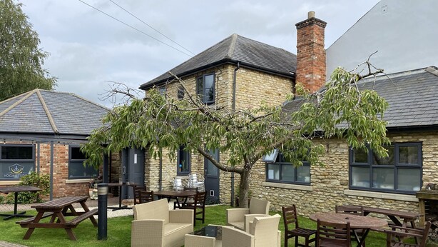 A stone building with a brick chimney, outdoor seating, and a tree. - The Roade House Rooms (Northampton)