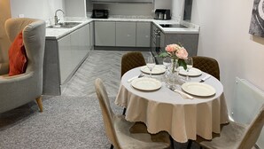 A modern kitchen with grey cabinets, a dining table set for two with white tablecloths, and a chair with a red cushion. - The Roade House Rooms (Northampton)