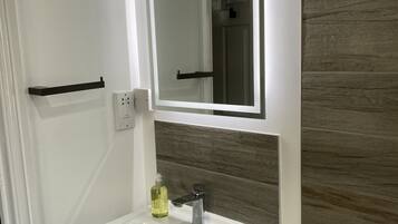 Un baño con un lavamanos blanco, un espejo y una pared de madera. Hay tres botellas de desinfectante para manos, cabello y cuerpo en la pared.