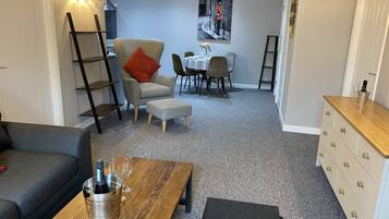 Ein gemütliches Wohnzimmer mit einer Couch, einem Holztisch mit einer Flasche und Gläsern, einem grauen Sessel mit einem roten Kissen, einem Esstisch mit Stühlen und einer Leiter an der Wand.