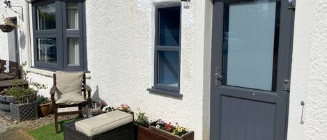 A white building with a grey door, a window, and a small outdoor seating area with a chair and cushions.