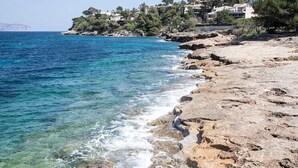Paisagem costeira com mar azul cristalino, margem rochosa e edifícios numa encosta.