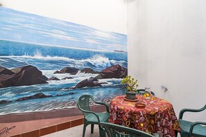 A table with a colorful tablecloth, a potted plant, and a glass of water on a balcony with a mural of the ocean.