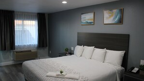Una cama bien hecha con edredón blanco y almohadas, una mesita de noche con lámpara y teléfono, y una ventana con cortinas.