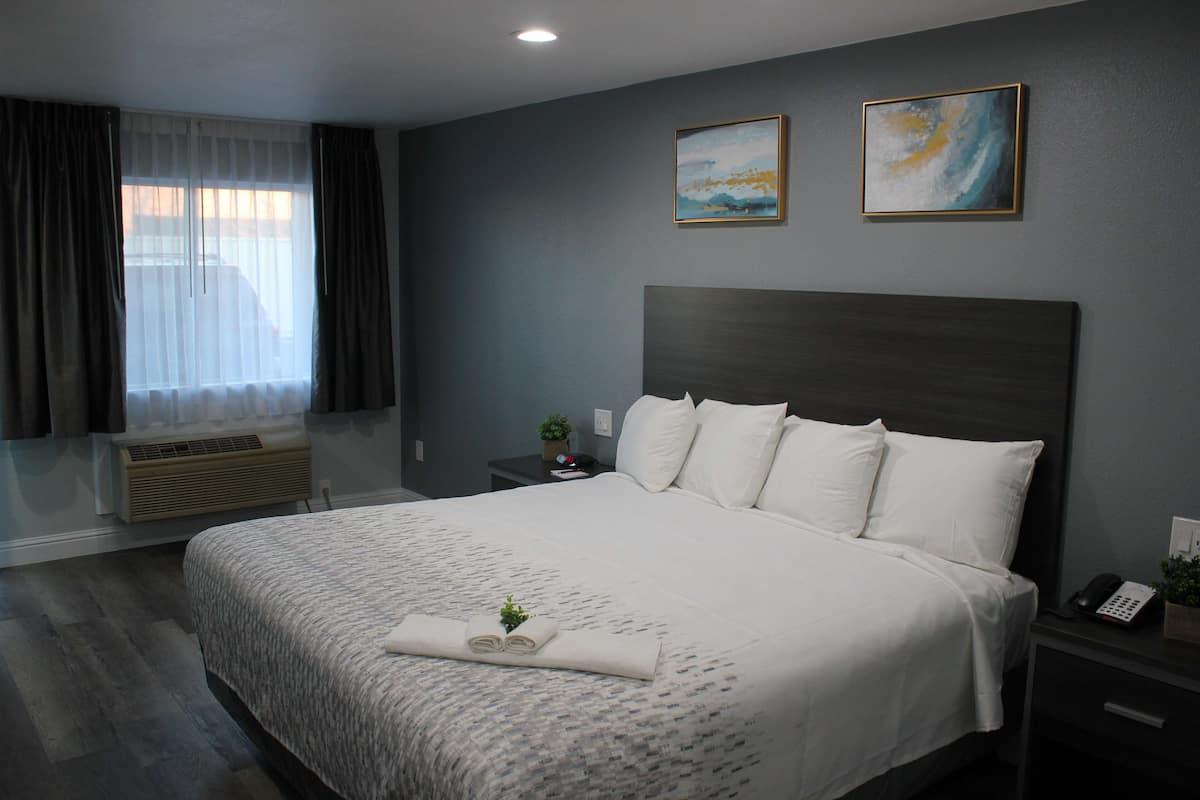 A neatly made bed with a white comforter and pillows, a bedside table with a lamp and a phone, and a window with curtains.