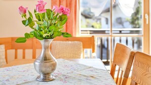 A table with a floral tablecloth, a silver vase with pink roses, and a window with a view of buildings. - Katharinenhof Holiday Apartment "Kramer Sehr Groß" with Balcony, Mountain View & Wi-Fi (Garmisch-Partenkirchen)