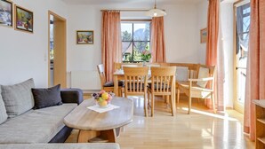 A living room with a sofa, a coffee table with a bowl of fruit, a dining table with chairs, and a window with curtains. - Katharinenhof Holiday Apartment "Kramer Sehr Groß" with Balcony, Mountain View & Wi-Fi (Garmisch-Partenkirchen)