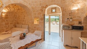 A stone-walled room with a bed, a table with a lace tablecloth, a basket of bread, and a kitchenette with a microwave. - Traditional Holiday Home Trullo Milù with WiFi, Air Conditioning (San Michele Salentino)