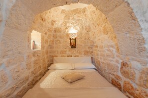 Une pièce aux murs de pierre, avec un lit, un luminaire fixé au mur et une petite fenêtre.