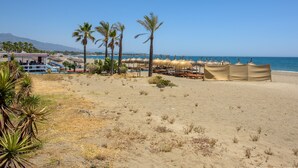 A beach with palm trees, beach umbrellas, and a building with a red roof. - Air-Conditioned Vacation Home in Villa Complex with Pool, Beach, Spa, Gym & Wi-Fi (Estepona)