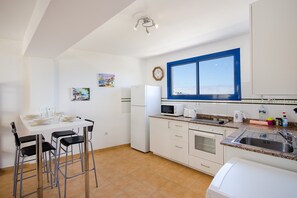 A kitchen with white cabinets, a white refrigerator, a microwave, and a sink. There are two bar stools and a dining table. - Charming Seafront Apartment with Balcony and Wi-Fi (Orzola)