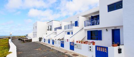 Une rangée de bâtiments blancs aux portes et fenêtres bleues, un chemin de gravier et un ciel dégagé.
