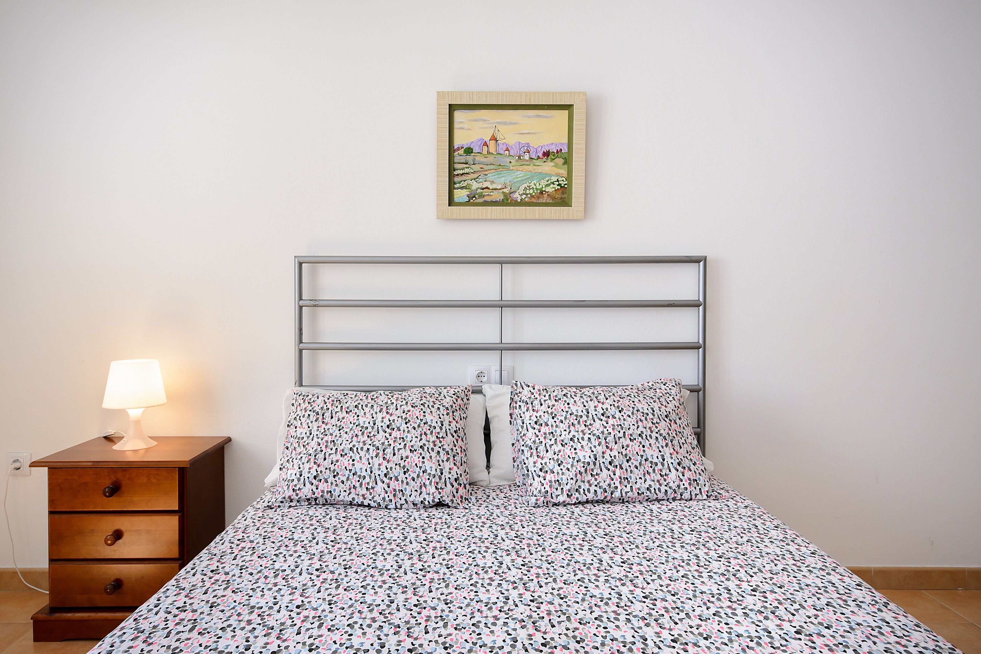 Un dormitorio con cabecera de metal, una mesita de noche de madera con lámpara, y una cama con ropa de cama estampada.