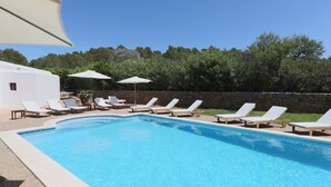 Una piscina con sillones de descanso y sombrillas, rodeada de un muro de piedra y árboles.
