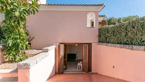Un bâtiment à la façade rose, doté d’une petite fenêtre en arc de cercle et d’une porte donnant sur un espace intérieur où se trouve une chaise.