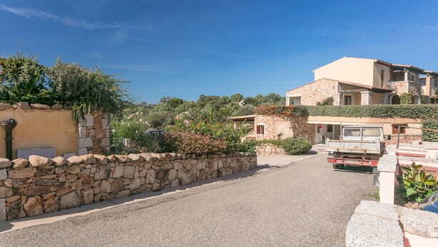 Un chemin bordé de murs en pierre mène à un bâtiment à deux étages, doté d’un toit plat et d’un balcon.