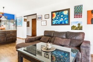 A living room with a brown leather sofa, a glass coffee table with a decorative tray, and a kitchen in the background.