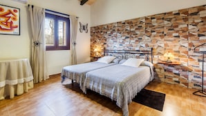 A bedroom with a stone wall, two beds, a window with curtains, a table, and a lamp. - Embedded in Nature and with Pool - Finca Esplendor (CAMPOS)