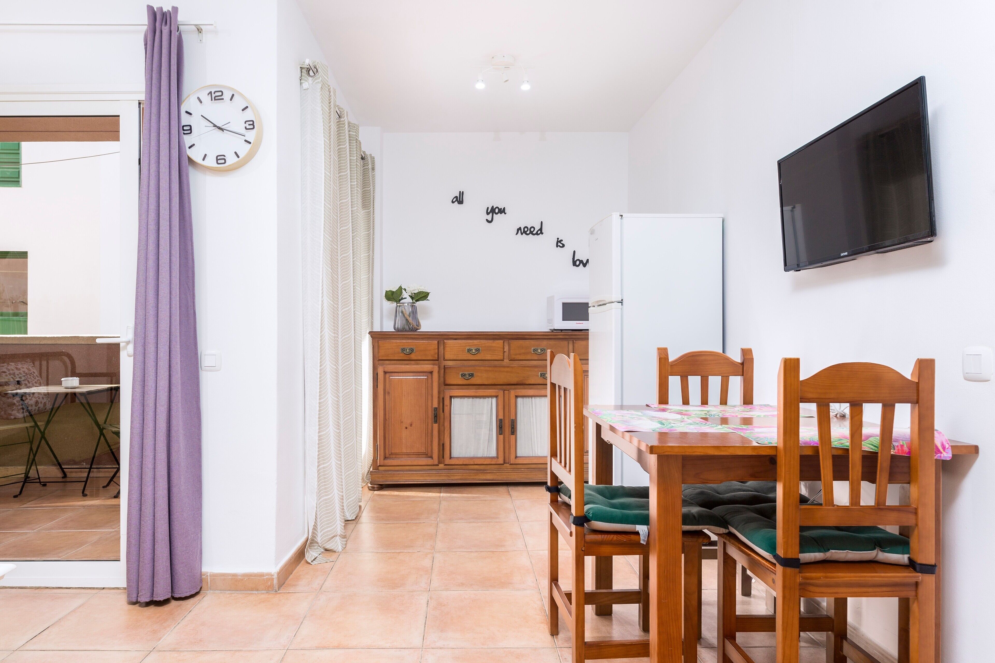 Un coin repas avec une table en bois, des chaises, une horloge, un téléviseur et, au fond, une cuisine.