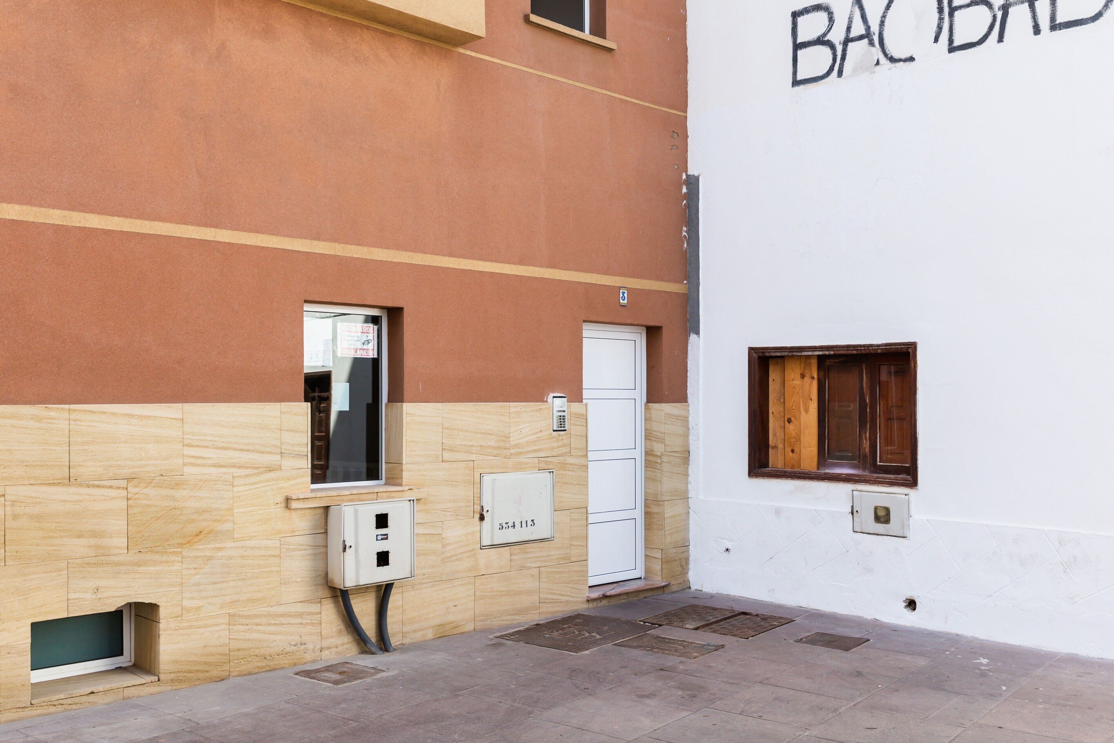 Un bâtiment à la façade beige et blanche, une porte avec une plaque de numéro, une fenêtre avec des volets en bois et un petit coffret électrique.