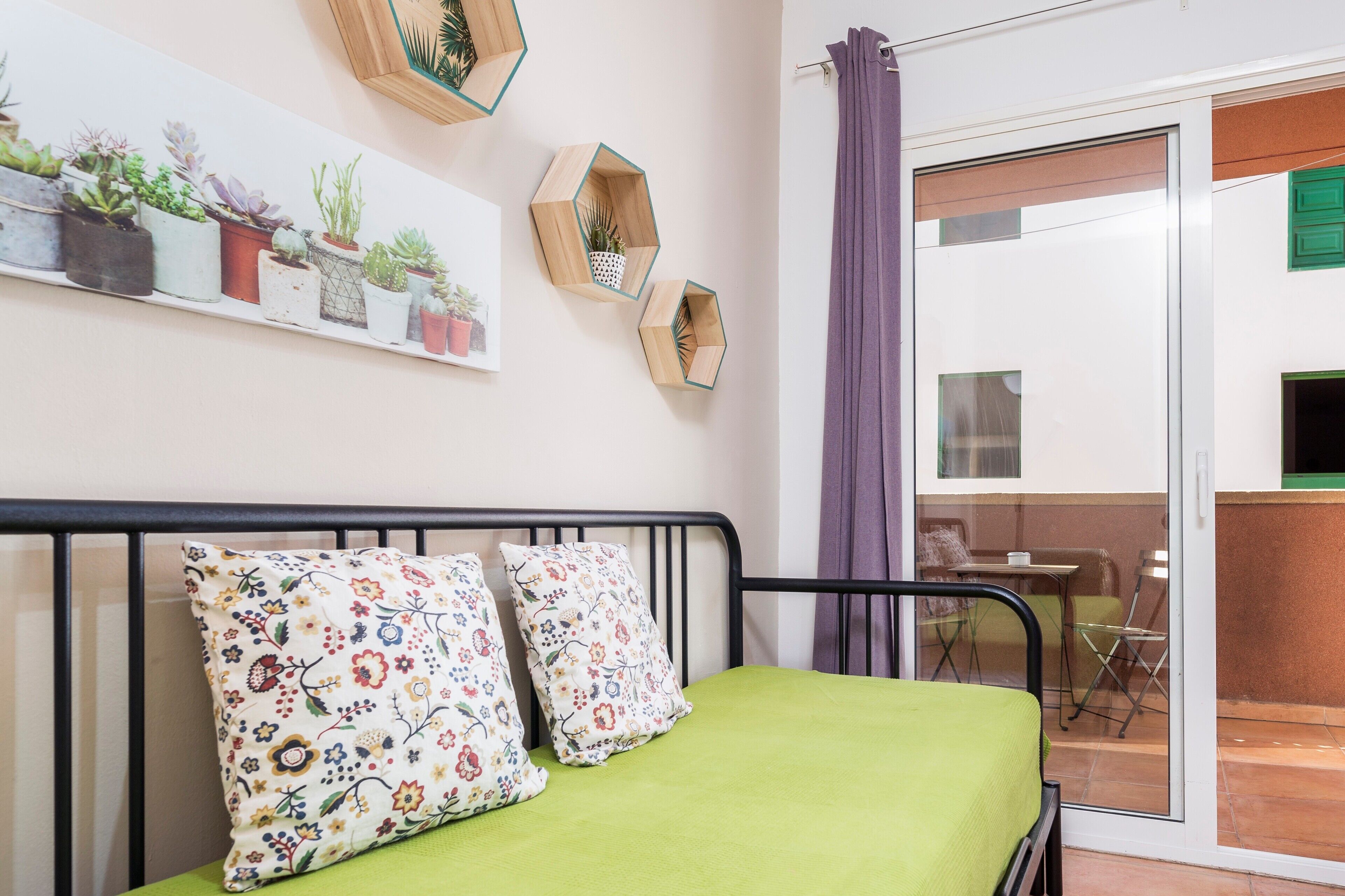 Un lit avec une literie verte, un coussin à motifs floraux, une étagère à plantes fixée au mur et une porte-fenêtre coulissante donnant sur un balcon.