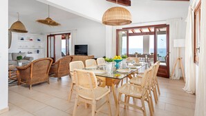Un comedor espacioso con sillas de mimbre y una mesa de vidrio, un área de estar con sofá y televisor, y vista al mar a través de amplias ventanas.