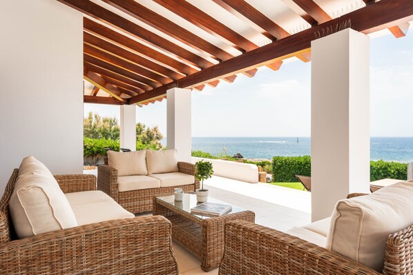 A covered outdoor seating area with wicker furniture, a glass coffee table, and a view of the sea.