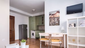 Ett kök med kylskåp, tvättmaskin och gröna skåp. Det finns en matplats med två stolar och ett bord, en tv som är monterad på väggen och en hylla med diverse föremål.