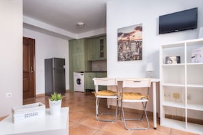 Une cuisine équipée d’un réfrigérateur, d’un lave-linge et de meubles de rangement verts. On y trouve un coin repas avec deux chaises et une table, un téléviseur fixé au mur et une étagère contenant divers objets.