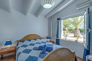 A bedroom with a bed, two bedside tables, a window with a view of palm trees and a building, and a ceiling fan.