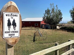 A welcome sign for Heart Strings Ranch with a red barn and a wooden fence in the background. - Heart Strings Ranch- Views of Heart Mountain & Near Horses! (Cody)