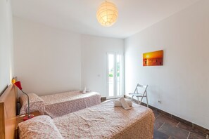 Une chambre avec deux lits, un bureau avec une lampe, une chaise, un tableau et une vue sur l’extérieur par une porte.