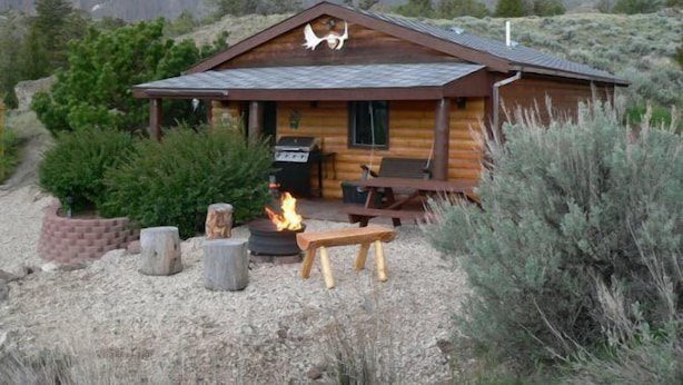 A rustic cabin with a chimney, a fire pit, and a bench.