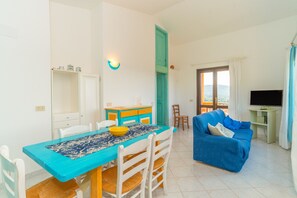 A living room with a blue sofa, a dining table with chairs, a TV stand, and a balcony with a view.