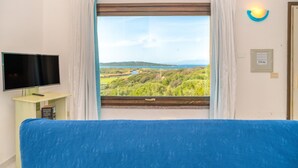 A room with a bed, a TV, a window with curtains, and a view of greenery and hills.