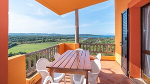 A balcony with a wooden table and white chairs, overlooking a green landscape and a body of water.