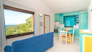 A room with a large window, a blue sofa, a kitchen with turquoise cabinets, and a dining area.
