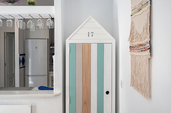 A room with a white wall, a wooden door with a number, a kitchen area with a refrigerator, and a macrame wall hanging.