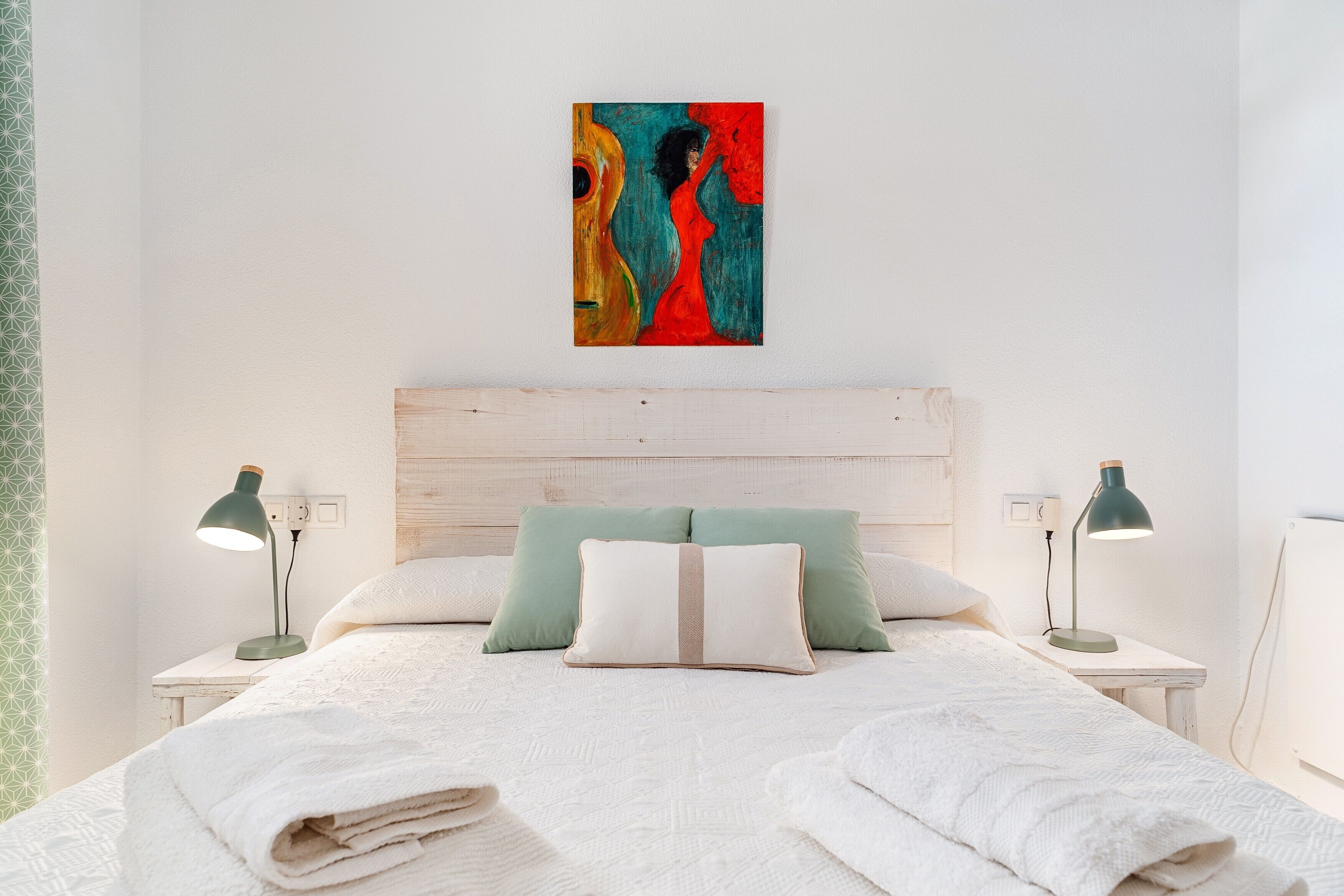 Ein ordentlich bezogenes Bett mit weißer Tagesdecke und zwei Kissen, ein Holz-Kopfstück und ein farbenfrohes abstraktes Bild an der Wand.