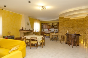 A living room with a yellow sofa, a dining table with chairs, a kitchen area with a stone wall, and a bar area with stools. - Fantastic Villa with Pool, Wi-Fi, Air Conditioning & Home Theater; Pets Allowed (Petacciato)