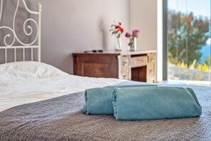 A neatly made bed with a grey blanket and two folded towels, a wooden nightstand with a vase of flowers, and a view of trees through a large window. - Air-Conditioned Villa with Sea View, Pool, Jacuzzi, Rooftop Gym/Terrace & Wi-Fi (Costa De La Calma, CALVIÀ)