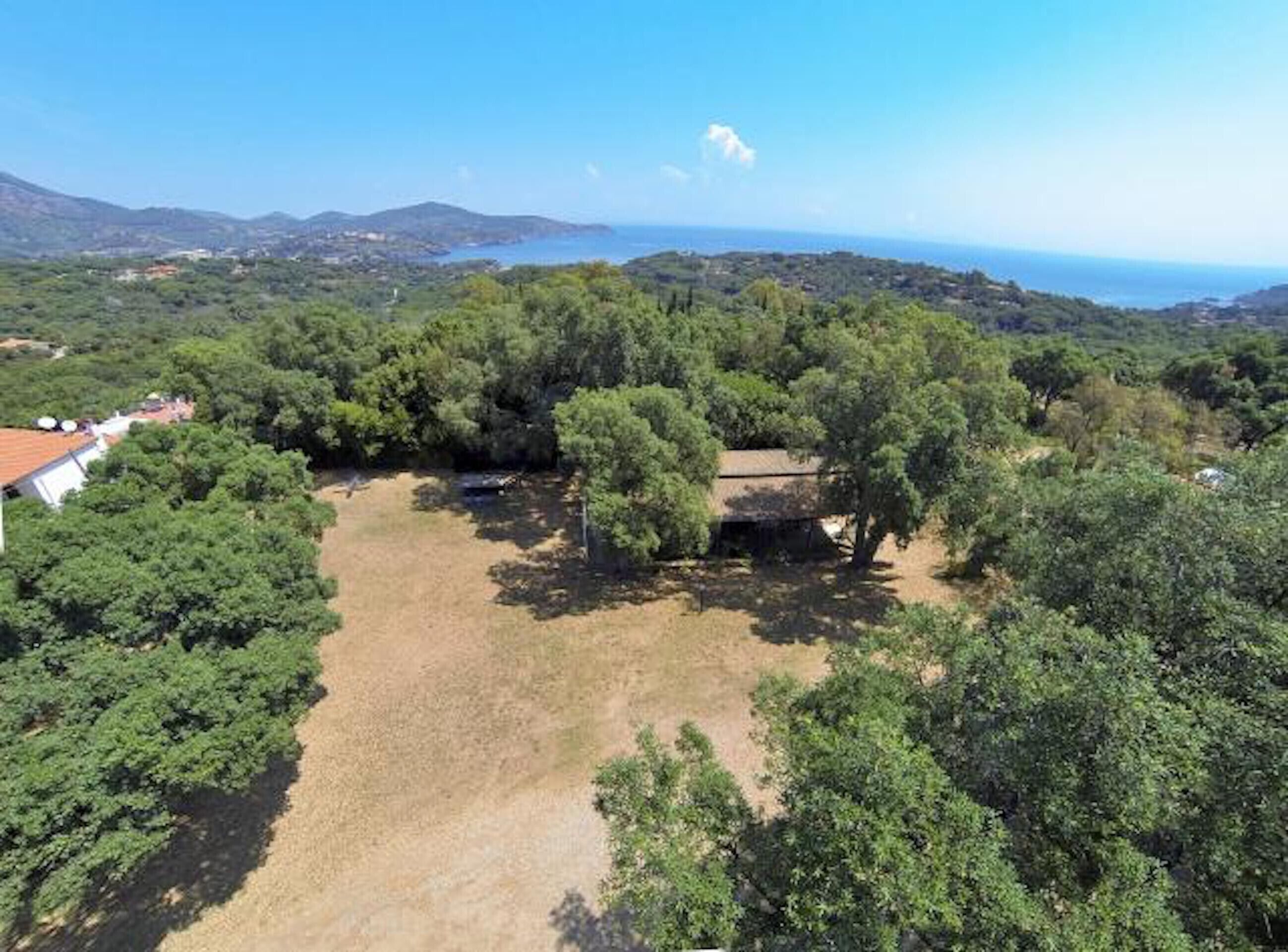 A rural property with a large open area, dense trees, and a view of the sea and hills in the background.