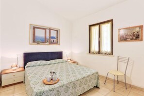 A bedroom with a bed, a chair, a nightstand, a window with curtains, and a wall-mounted picture.