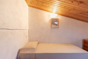 Una camera da letto con soffitto in legno, un letto con materasso bianco, un piccolo specchio a parete e un comò in legno.