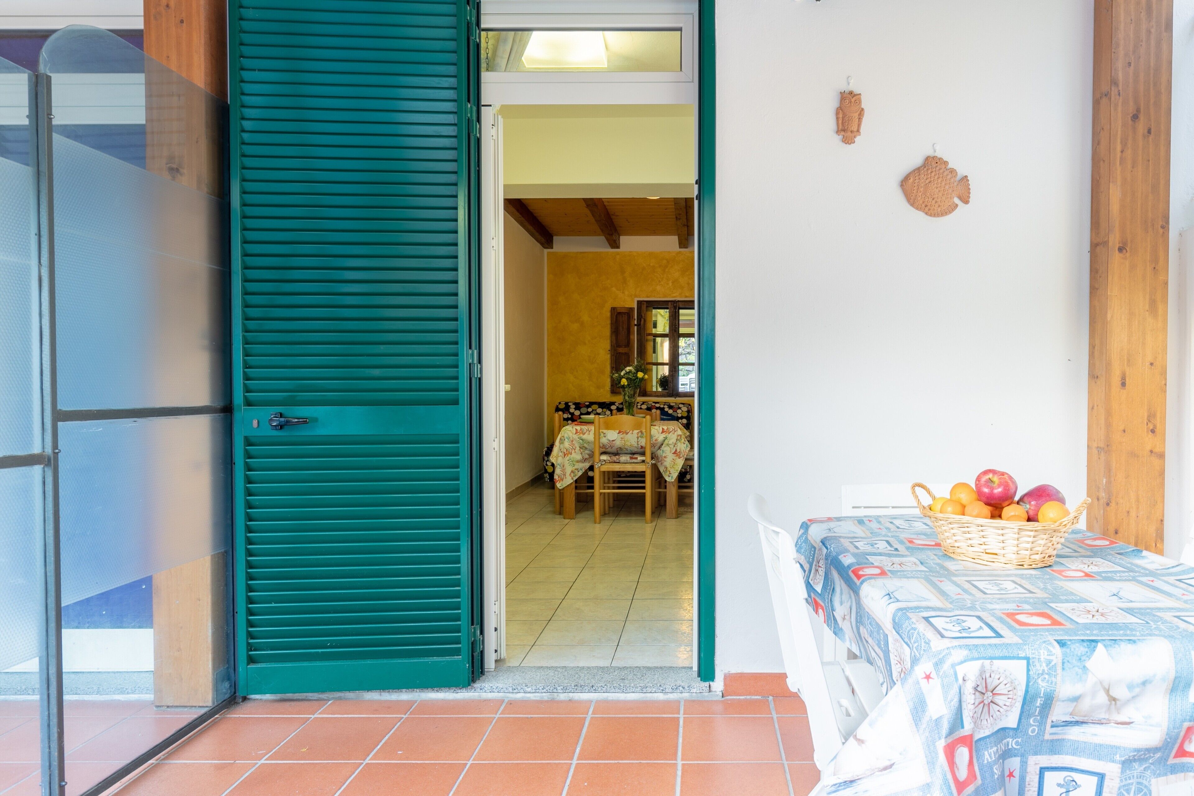 A room with a green shutter door, a dining area with a table and chairs, and a basket of fruit on a table.