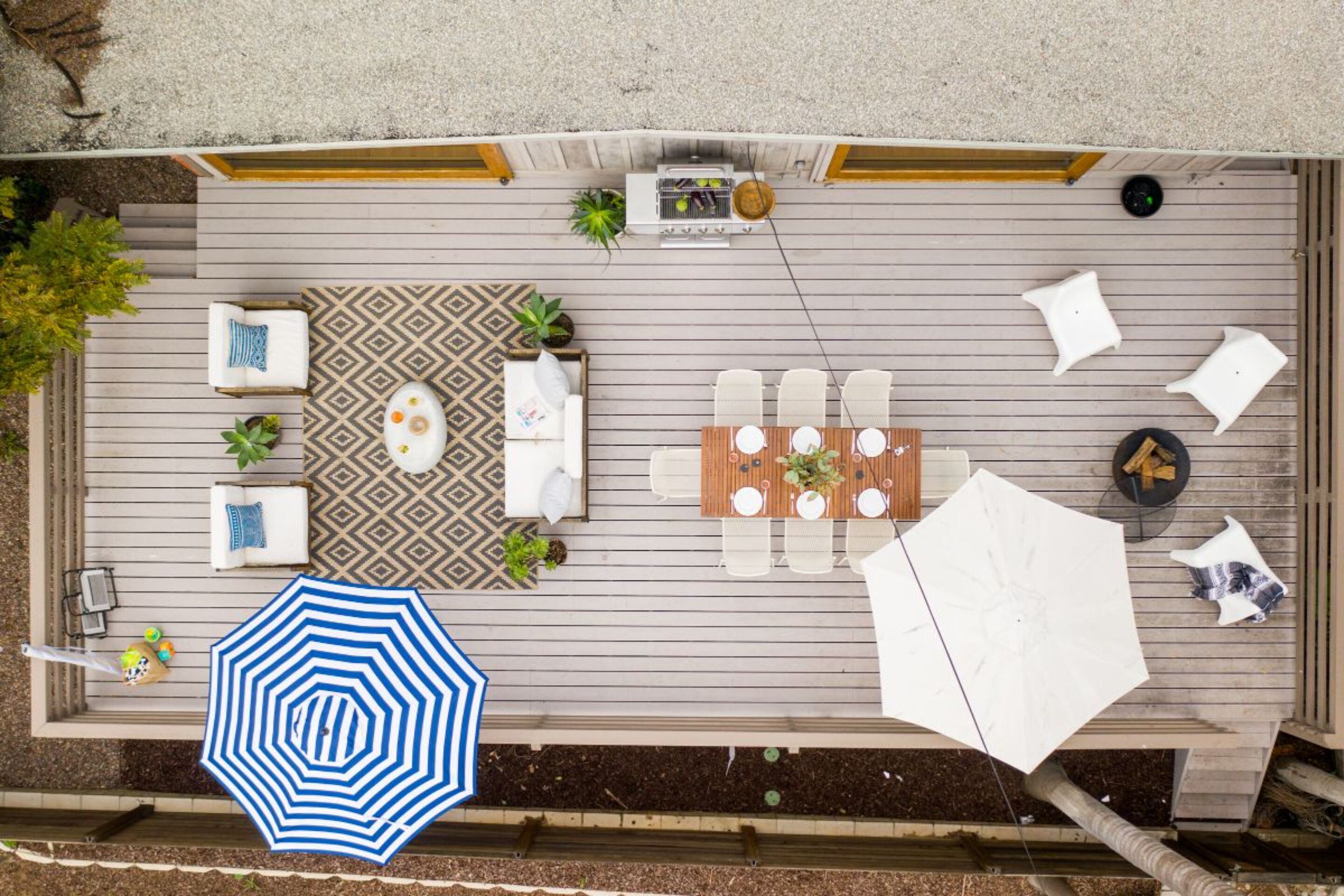 Een houten terras met buitenmeubilair, waaronder een tafel, stoelen, een gekleurd vloerkleed en een blauw-wit gestreepte parasol.