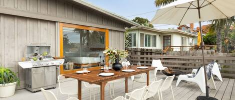 An outdoor patio with a wooden table set for dining, surrounded by white chairs, a grill, and a large umbrella.