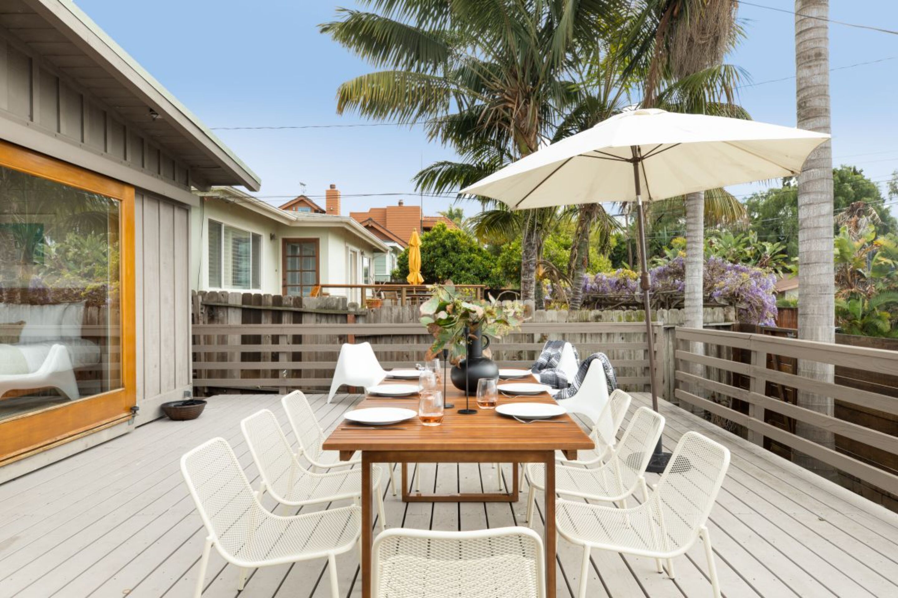 Une terrasse en bois avec une table dressée pour quatre personnes, entourée de chaises blanches, et un grand parasol qui offre de l’ombre.